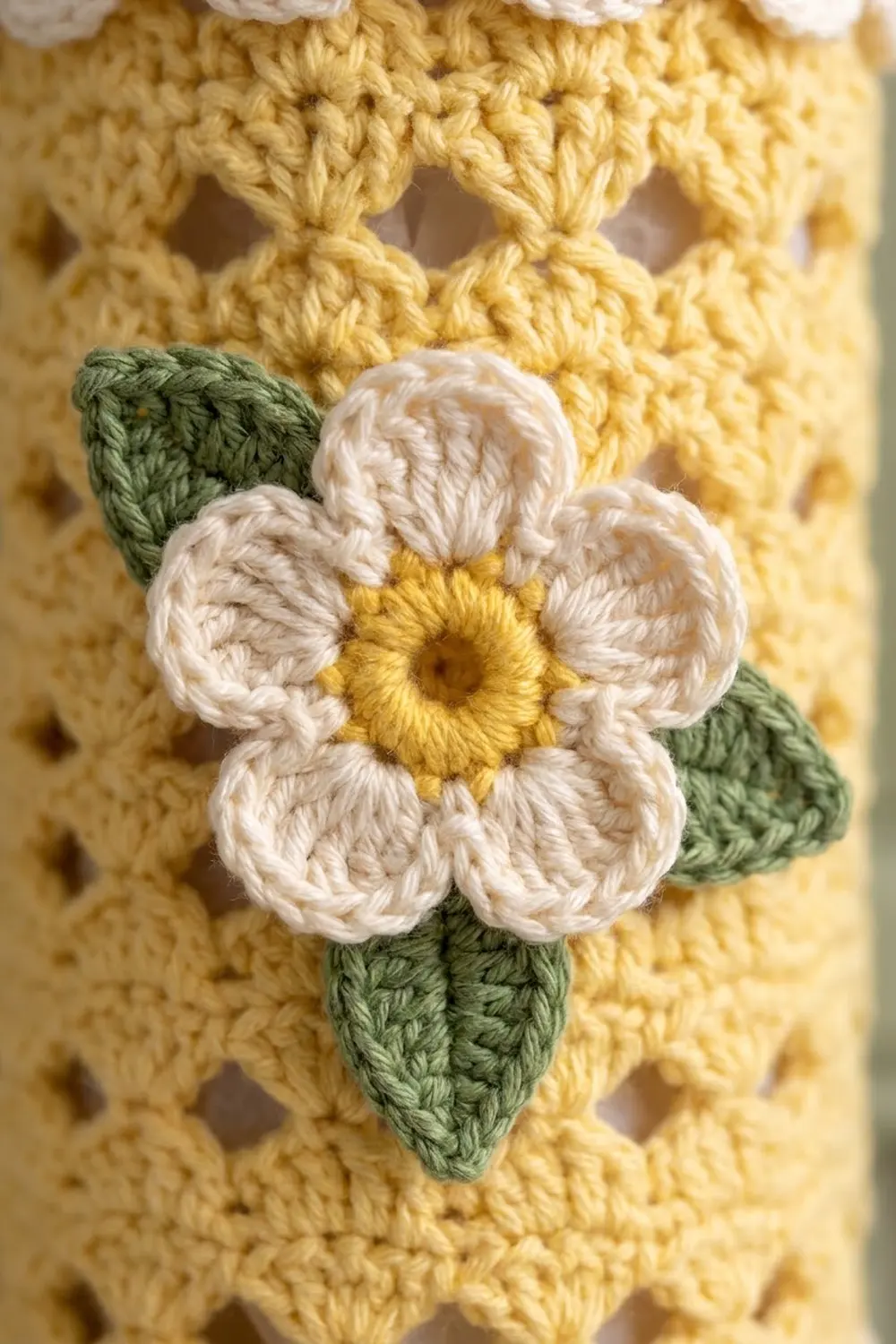 White crocheted flower with yellow center and green leaves appliqued on pale yellow knit fabric.