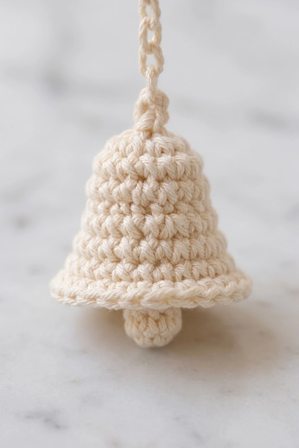 Cream crocheted bell ornament with detailed stitch texture hanging from braided cord against neutral background.