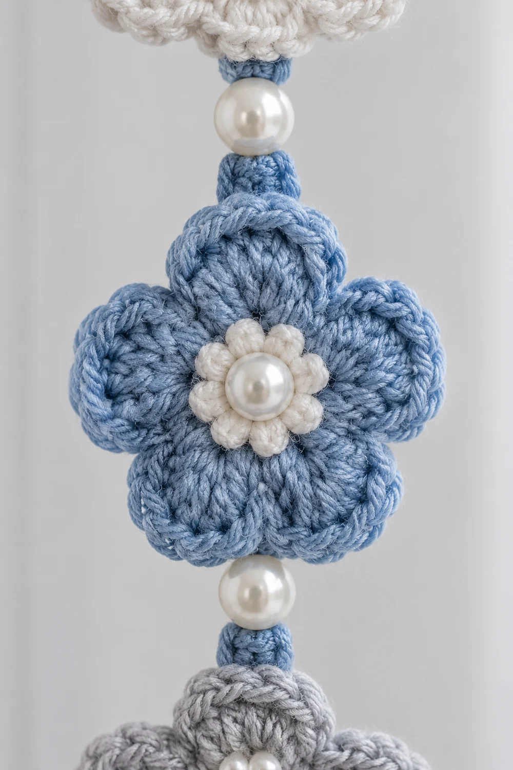 Crocheted blue flower with white pearl center suspended between pearl beads on a light background.
