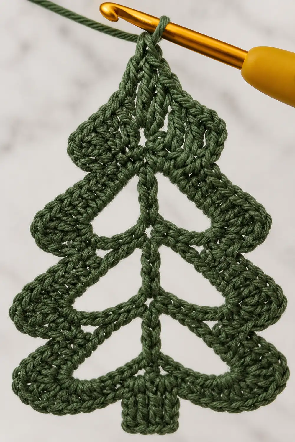 A crocheted green Christmas tree ornament hanging from a golden crochet hook against a white background.