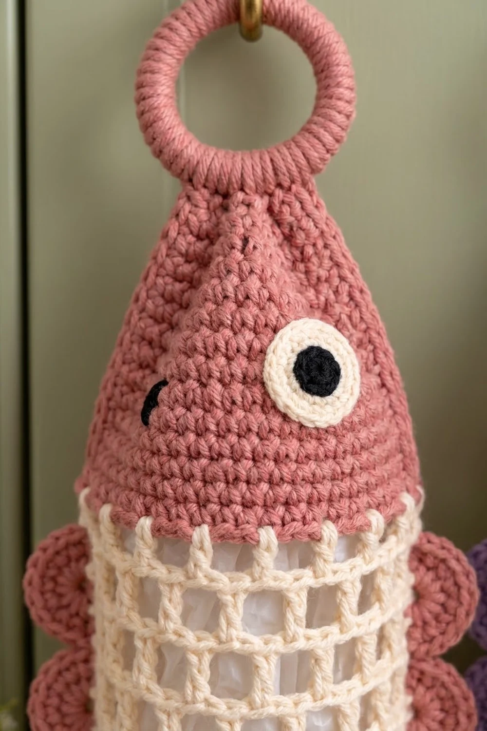 Close-up of a pink crocheted creature with white gridded base, embroidered eye, and brass ring handle.