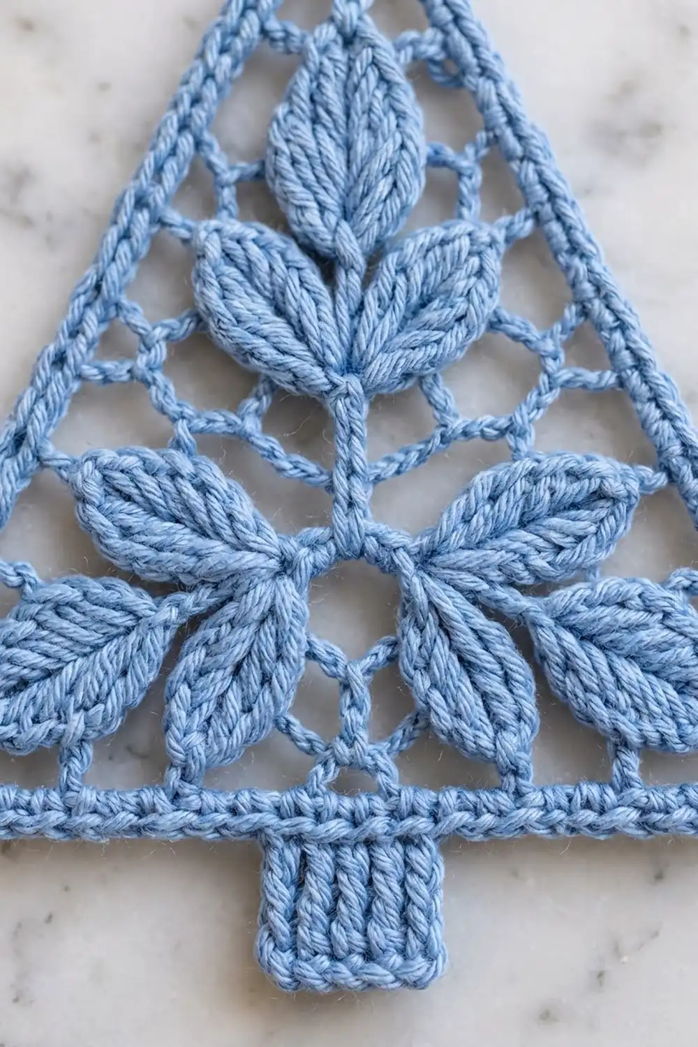 Light blue crocheted Christmas tree with textured leaf appliqués on a mottled marble background.