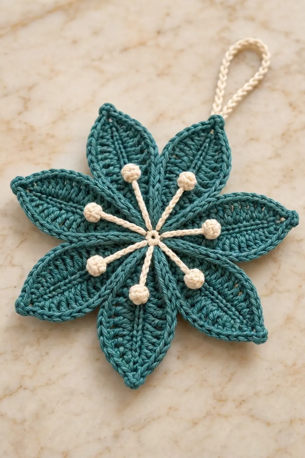 Handmade teal crochet flower with cream beaded stamens and loop, on marble surface.