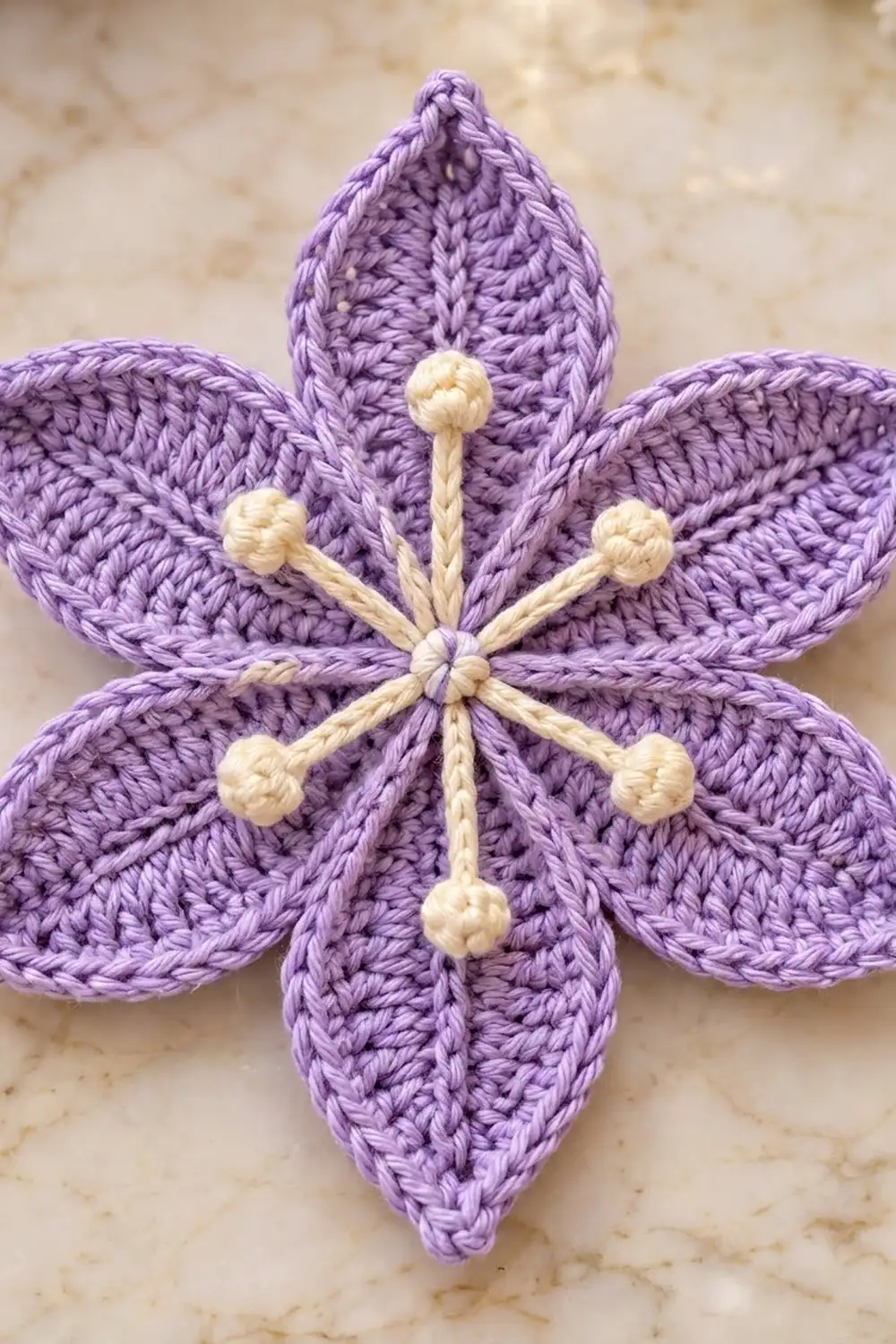 Purple crocheted five-petal flower with cream bobble-topped stamens on a neutral beige textured background.