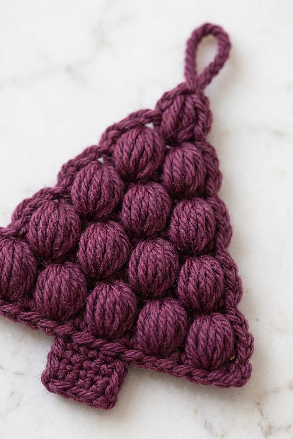 Plum crocheted pompon with braided hanging loop displayed on white marble.