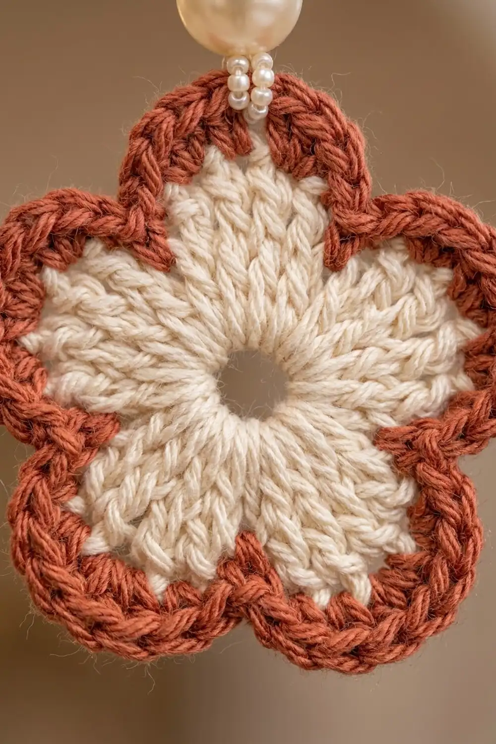Close-up of a crocheted flower ornament featuring cream textured center and rust-red petals with pearl bead hanging loop.