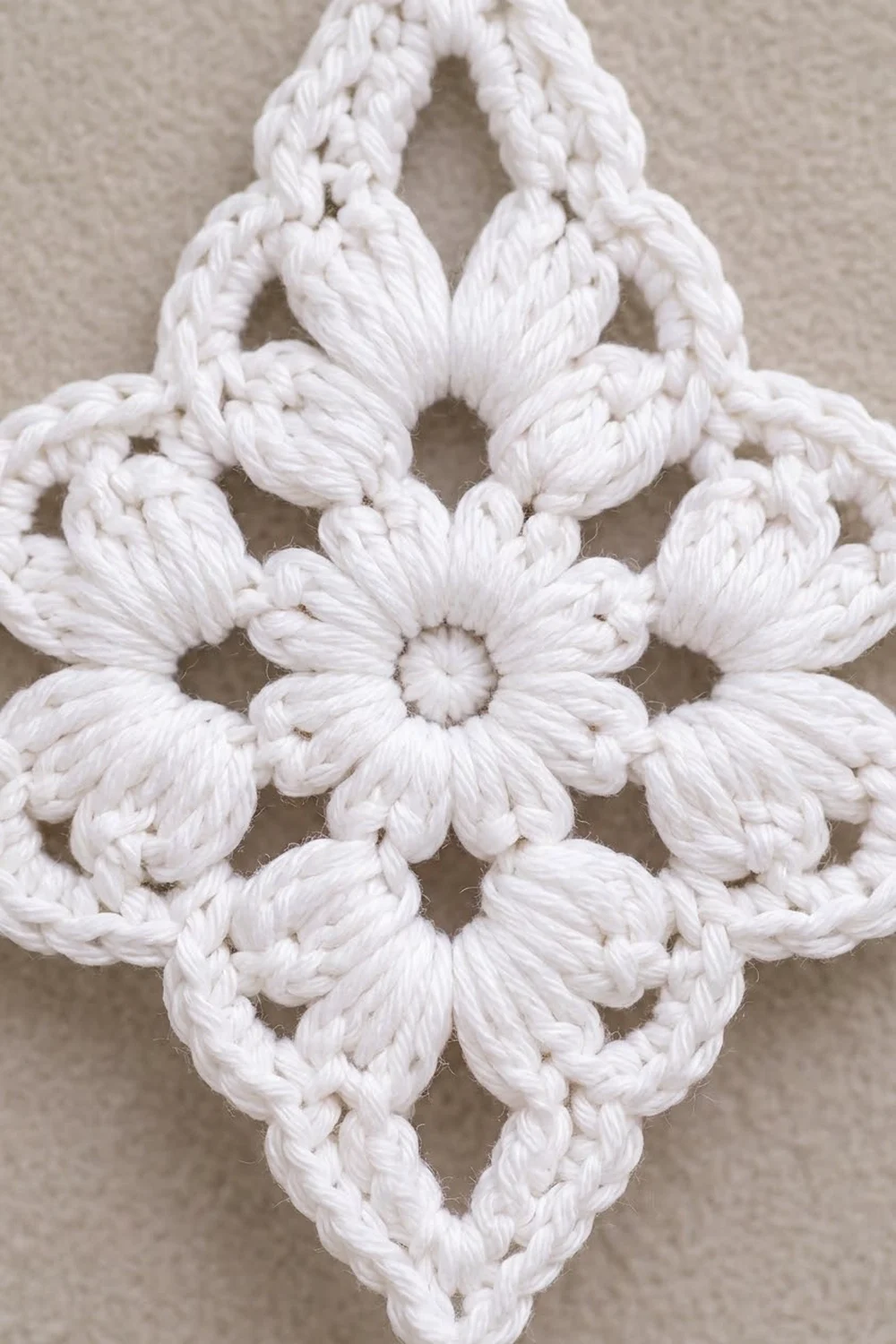 White crocheted motif with rope-textured petals arranged in a symmetrical pattern against neutral beige fabric.