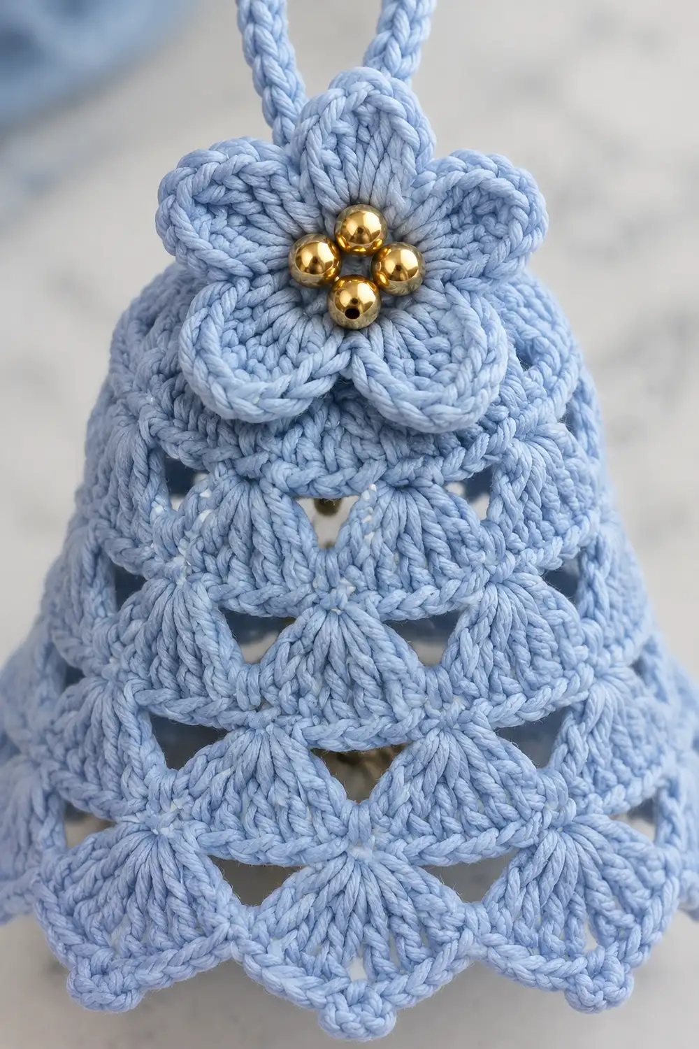 A light blue crocheted bell ornament with golden beads adorning its flower-shaped top and braided cord.