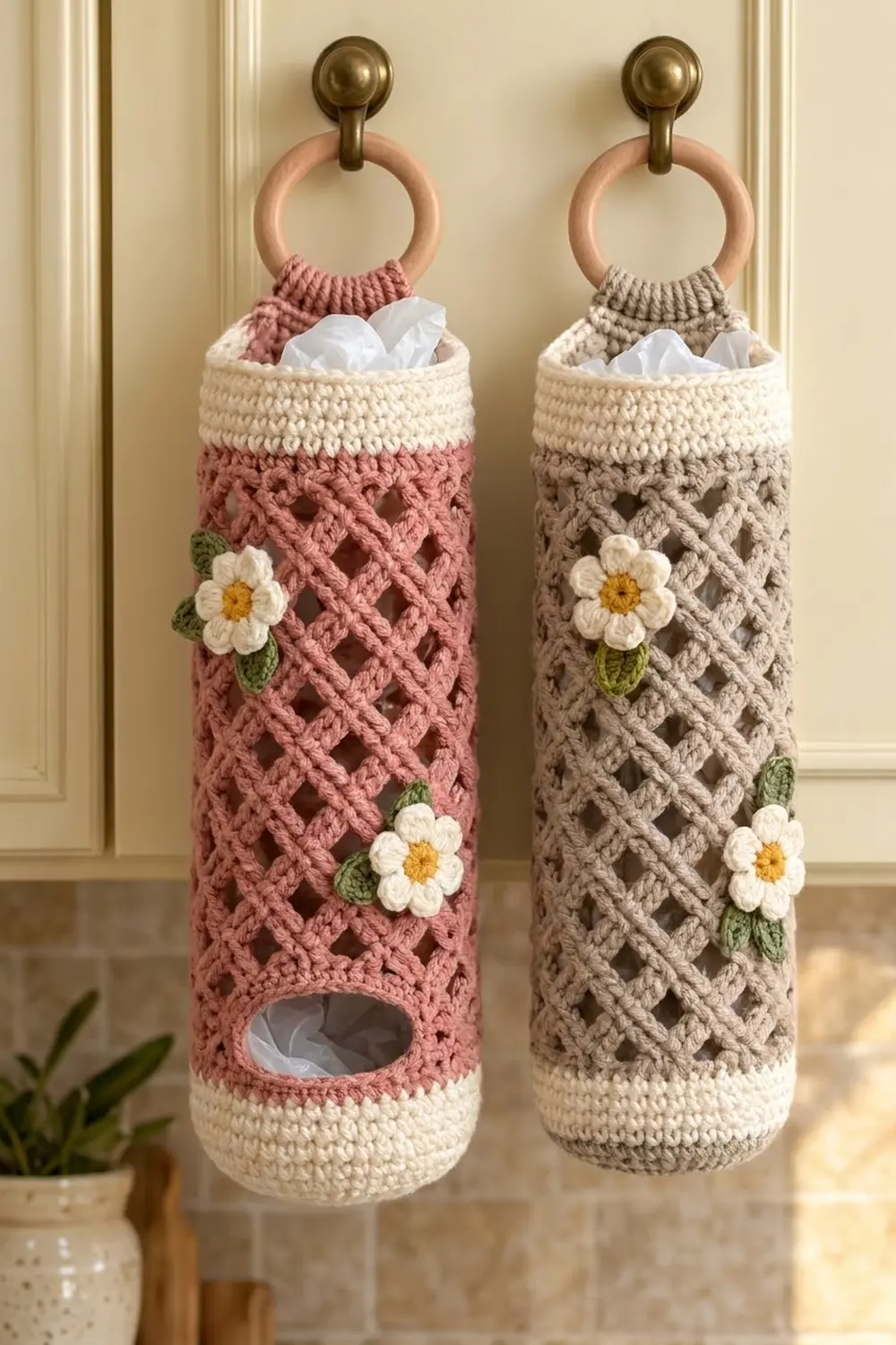 Two crocheted tissue holders in pink and gray with white daisies hang from brass finials on a pale wall.