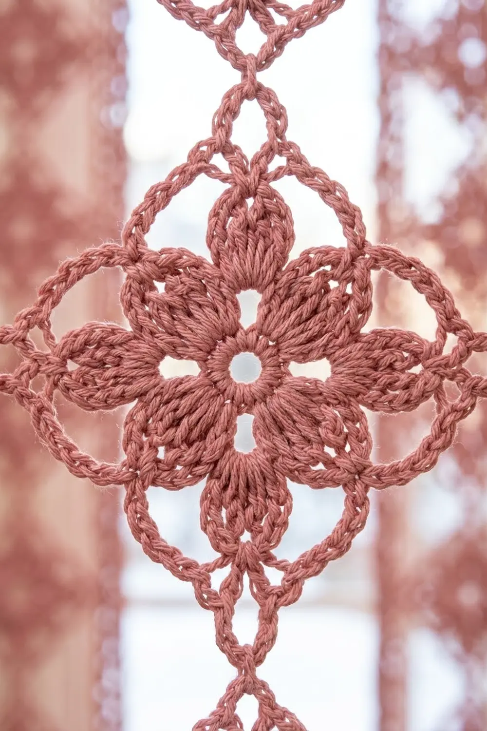 Delicate dusty rose crocheted ornament with intricate knotwork forming a symmetrical cross pattern against blurred pink and white background.