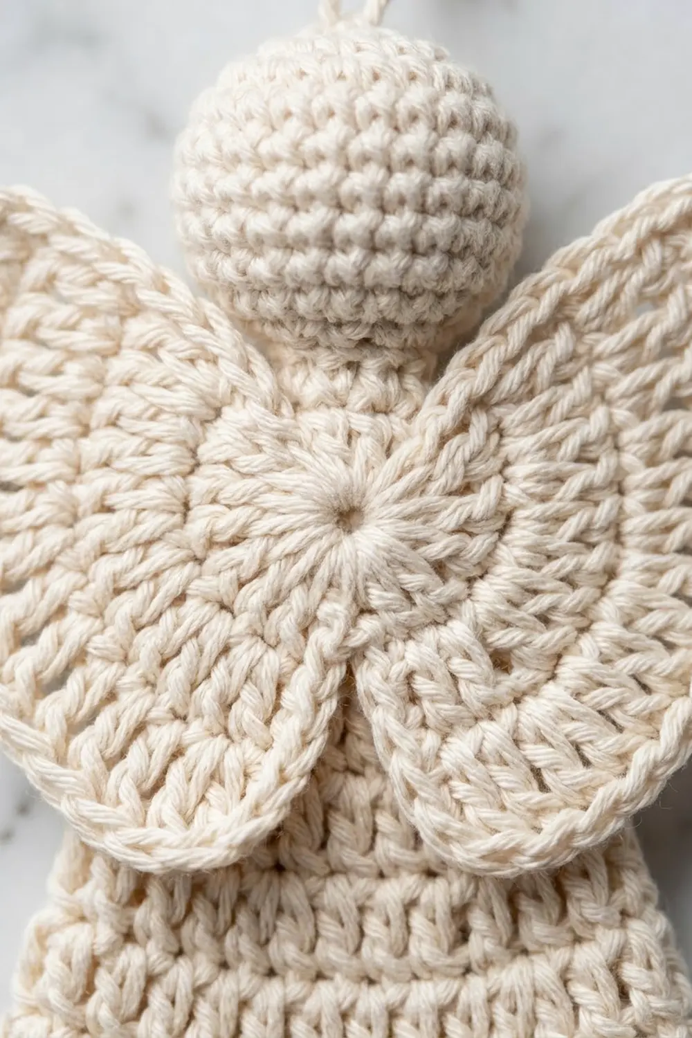Close-up of a cream-colored crocheted angel or fairy with a spherical head and delicate wings against a soft white background.
