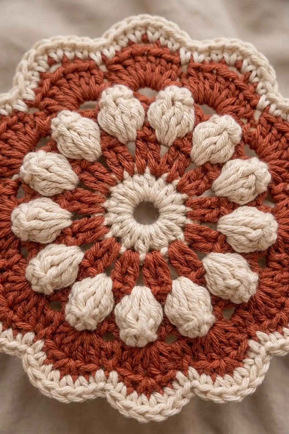 Crocheted square in terracotta and cream features a dimensional flower design with textured petals, set against a soft blurred background.