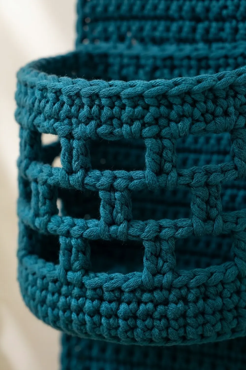 Teal crocheted fabric with geometric openwork pattern showing chunky textured stitches and square-shaped holes in detailed close-up.