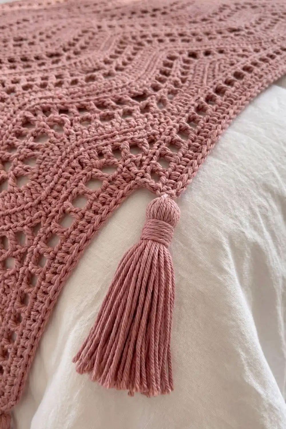 Dusty rose crochet blanket with geometric lace stitches and matching tassel trim draped over white fabric.