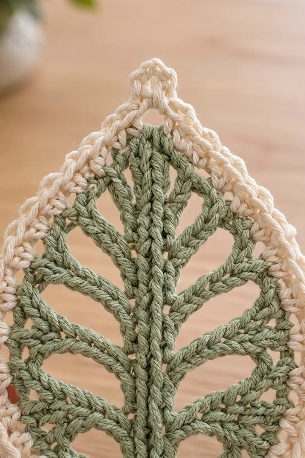 Close-up of crocheted leaf pattern in sage green yarn with cream-colored border stitches on a neutral background.