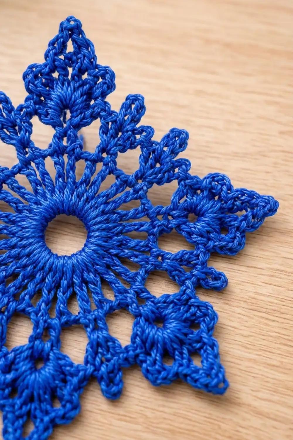 Intricate blue crocheted snowflake with detailed lacey openwork pattern resting on a natural wood background.