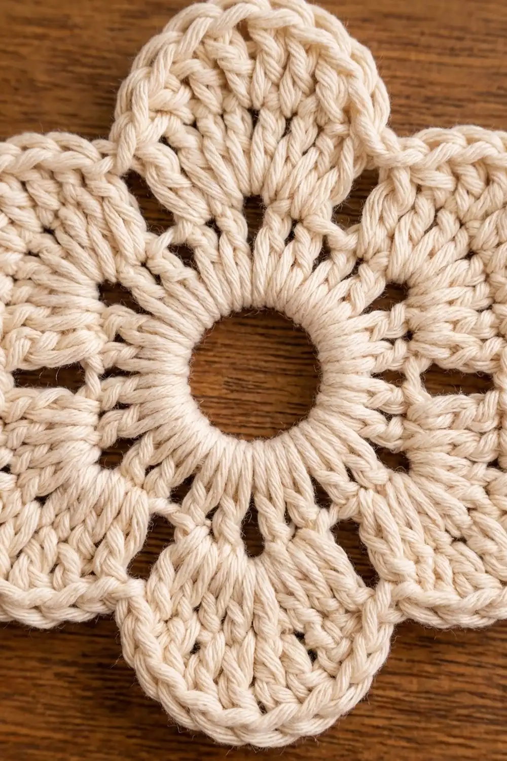 Ivory crocheted flower with four twisted petals and central hole on rich brown wooden surface.