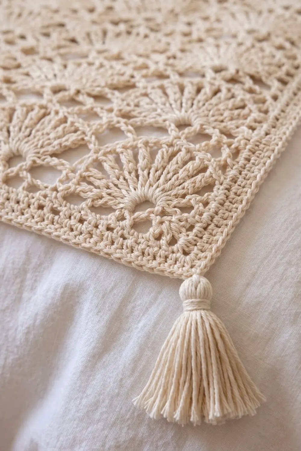 Cream-colored crocheted lace detail with geometric open pattern and decorative tassel against a neutral fabric background.