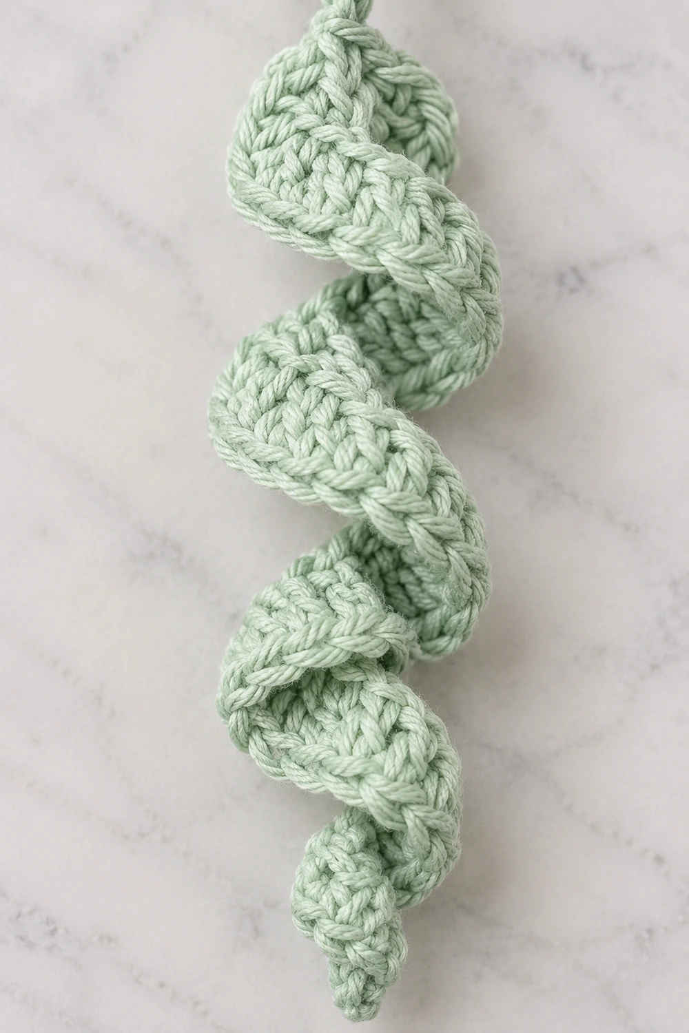 A coiled green crochet rope with textured braided pattern lies on a white marble surface.