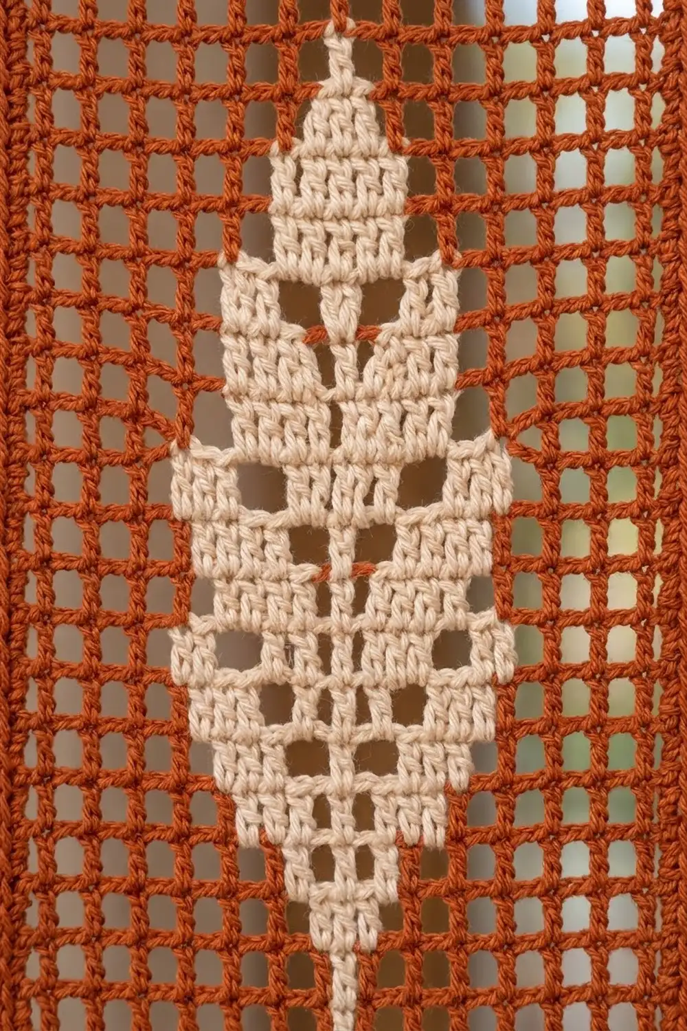 Cream-colored crochet cross with openwork filet stitches centered on a rust-orange geometric lattice pattern.
