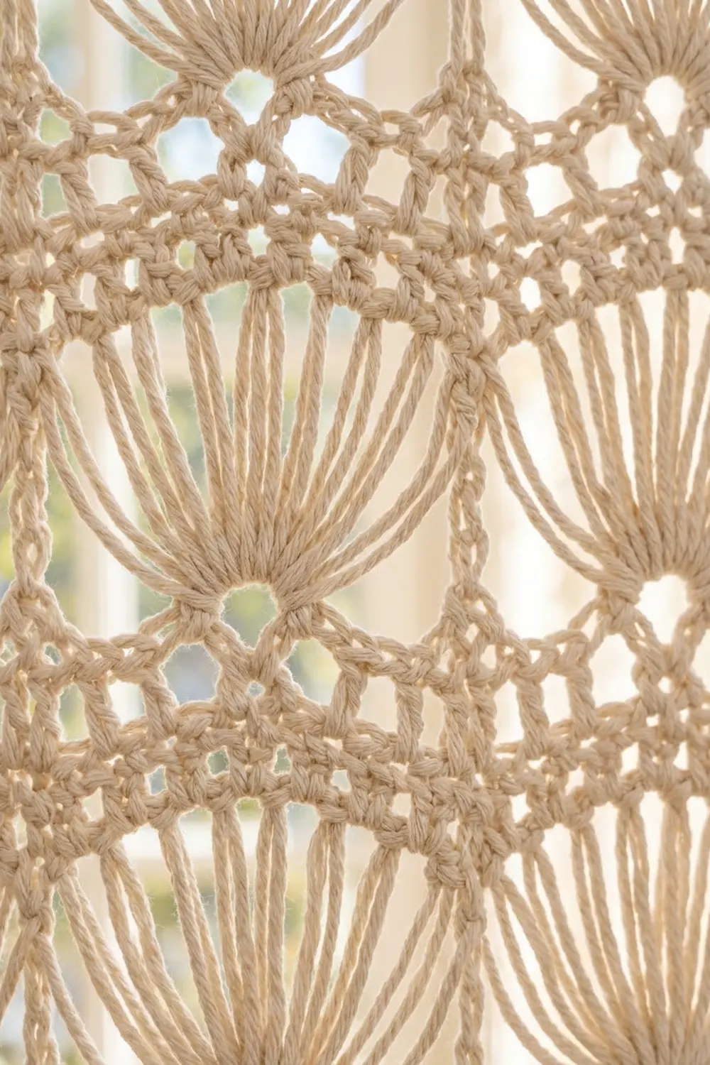 Cream-colored macramé with knotted geometric patterns and hanging fringe, illuminated by soft backlight.