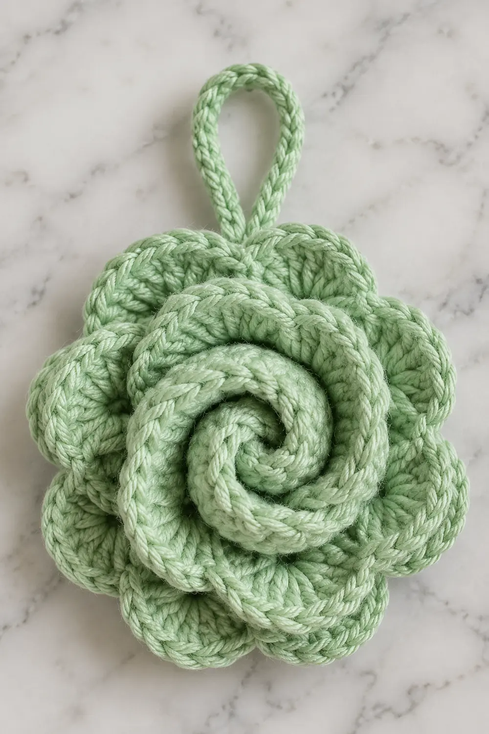 A pale green crocheted flower ornament with a spiral center and hanging loop on marble background.