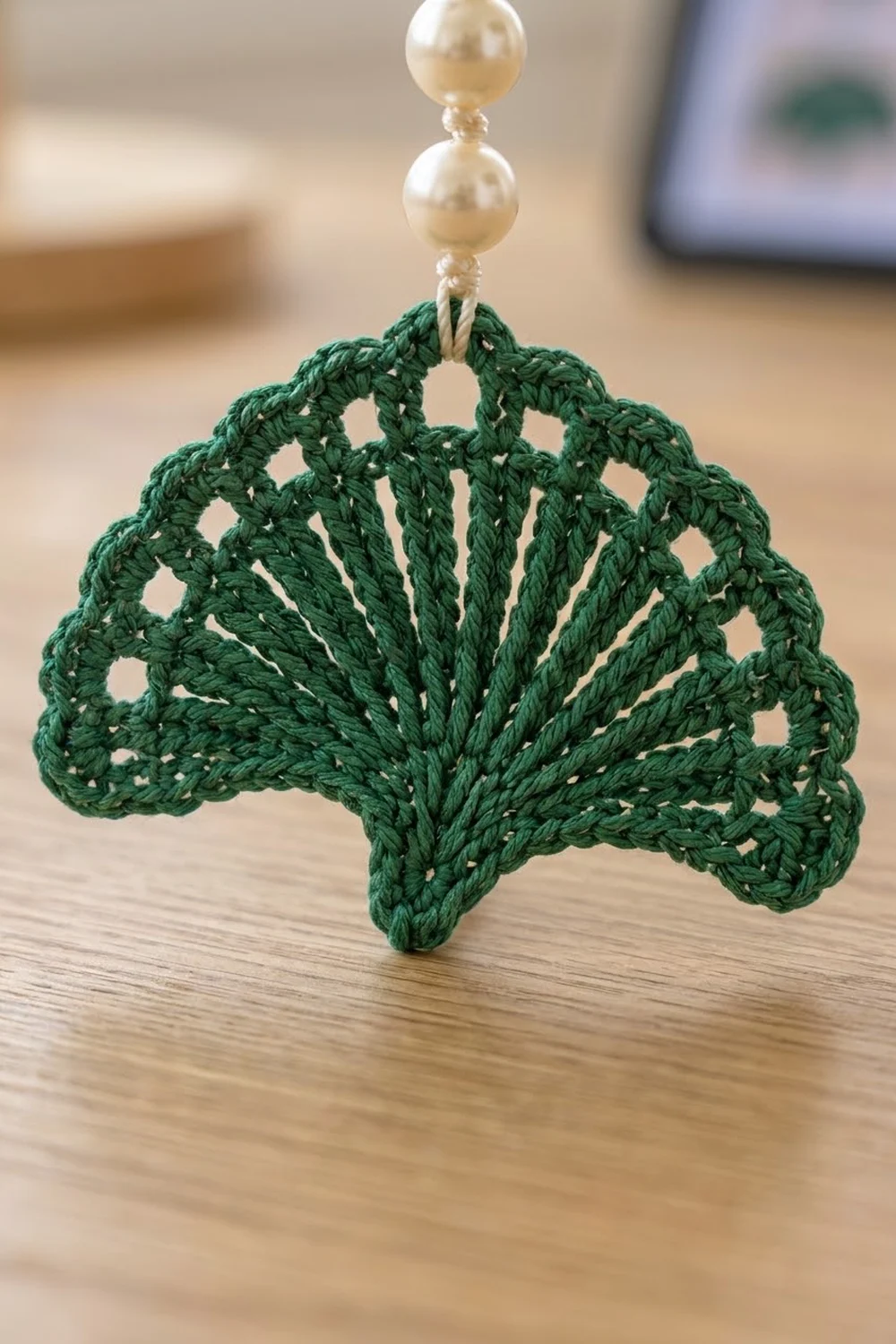 Dark green crocheted shell ornament with delicate openwork hangs from cream pearls on wood.