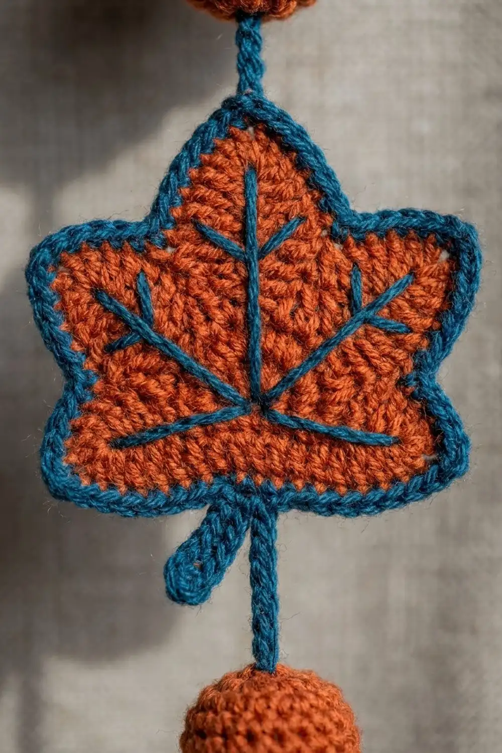 Crocheted maple leaf ornament with burnt orange sections and teal outline, hanging against a blurred white background.