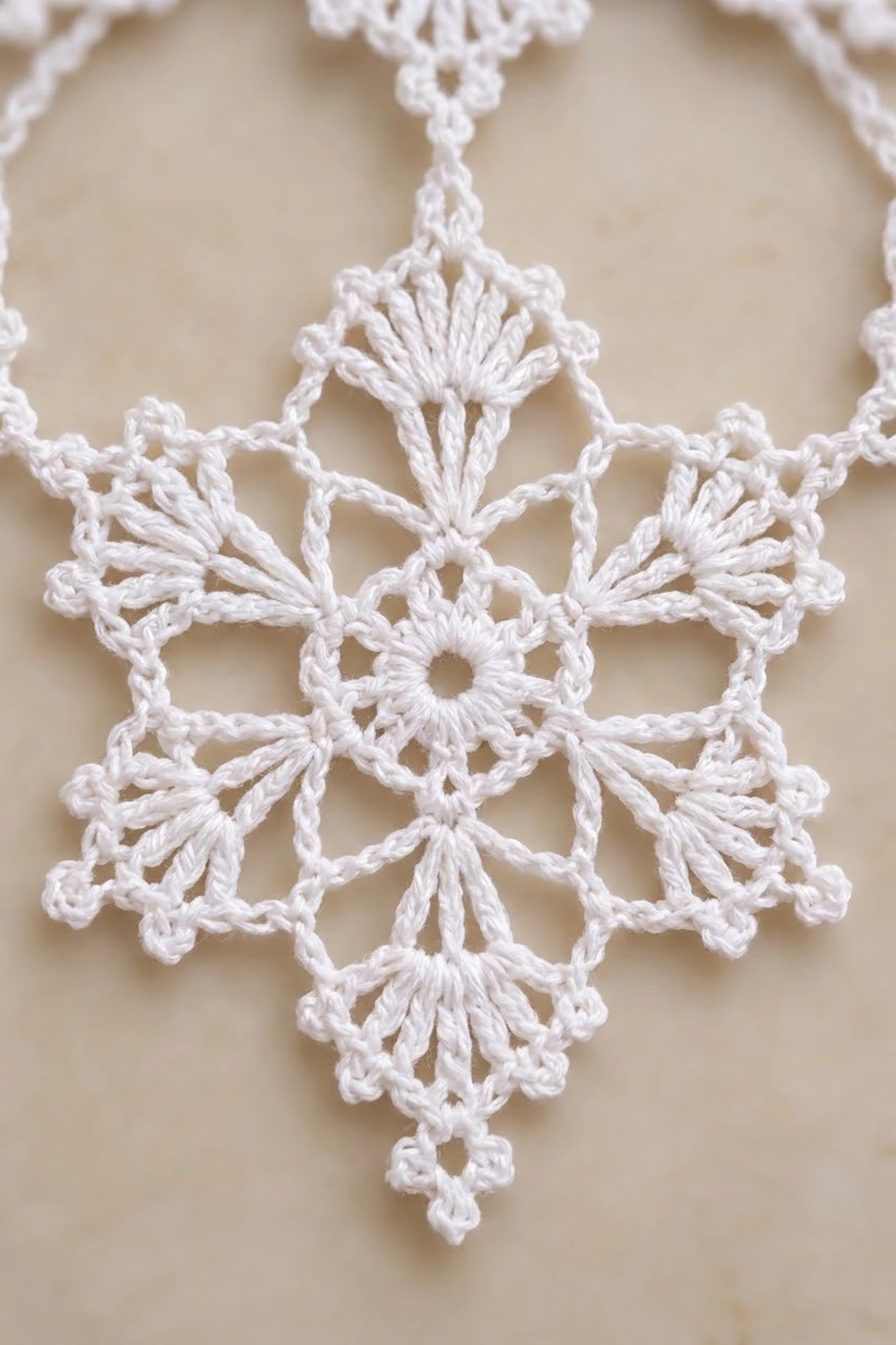 A white crocheted snowflake ornament with delicate lace fan patterns against a cream-colored background.
