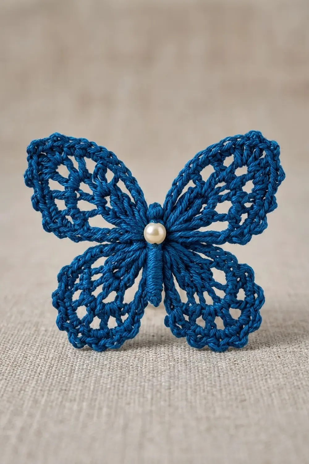 Bright cobalt-blue crocheted butterfly with white pearl bead center displayed on soft neutral linen fabric.