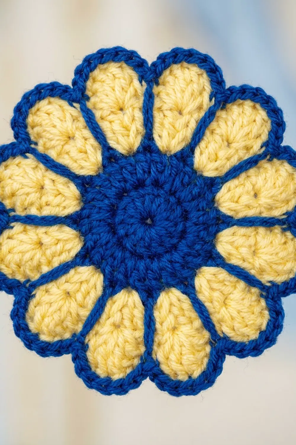Crocheted flower with twelve pale yellow petals edged in bright blue yarn, showing detailed stitch texture on a neutral background.