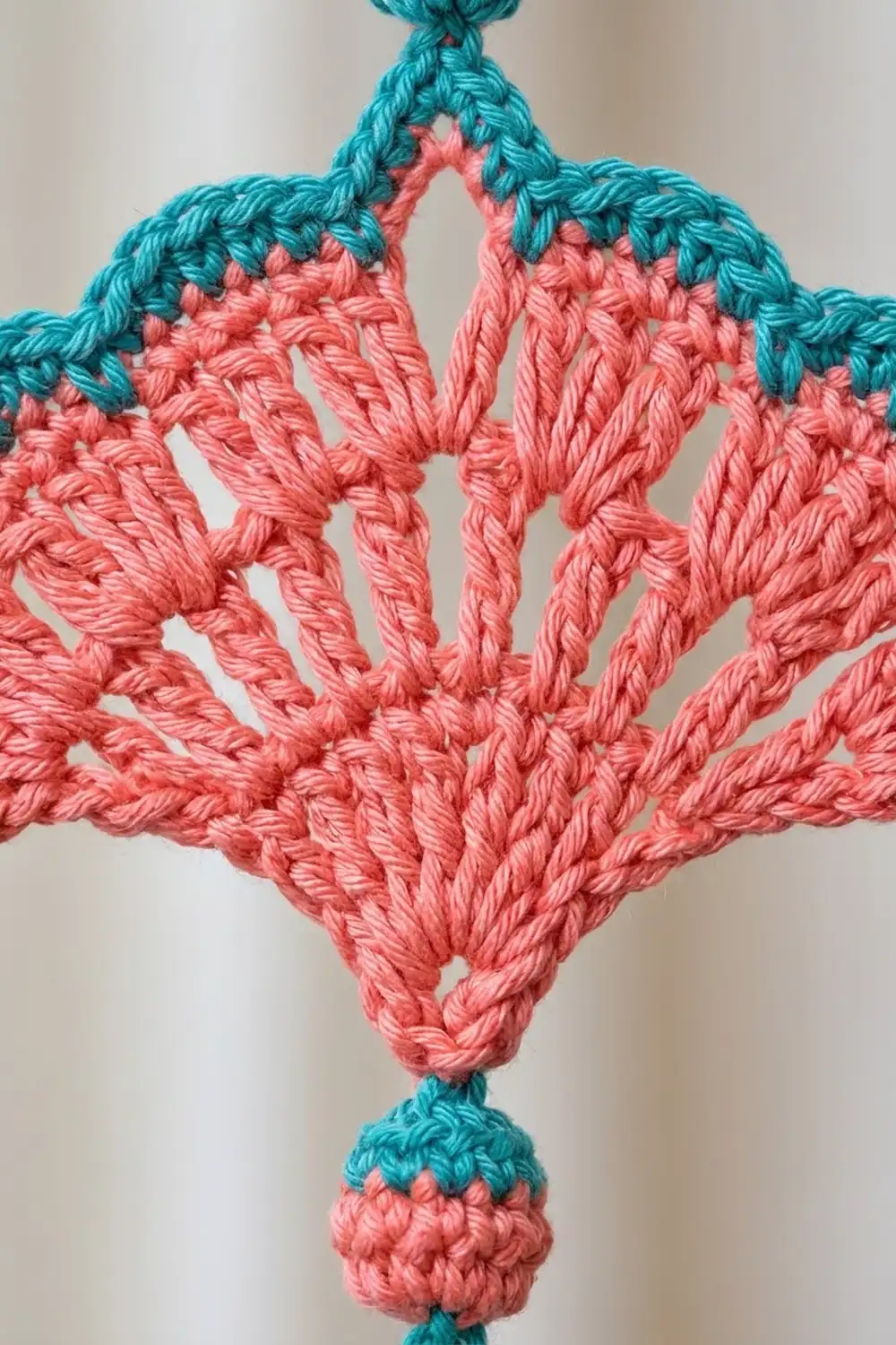 Crocheted diamond ornament in coral pink with teal borders, hanging against a neutral cream background.