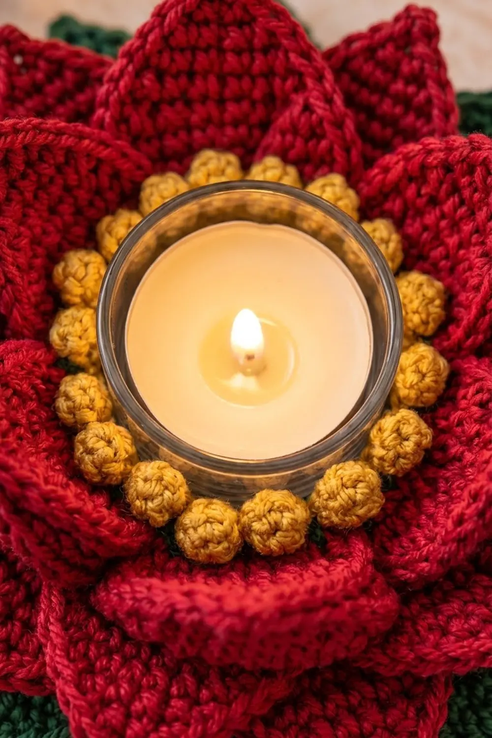 A lit candle in a glass holder surrounded by a red and gold crocheted flower design.