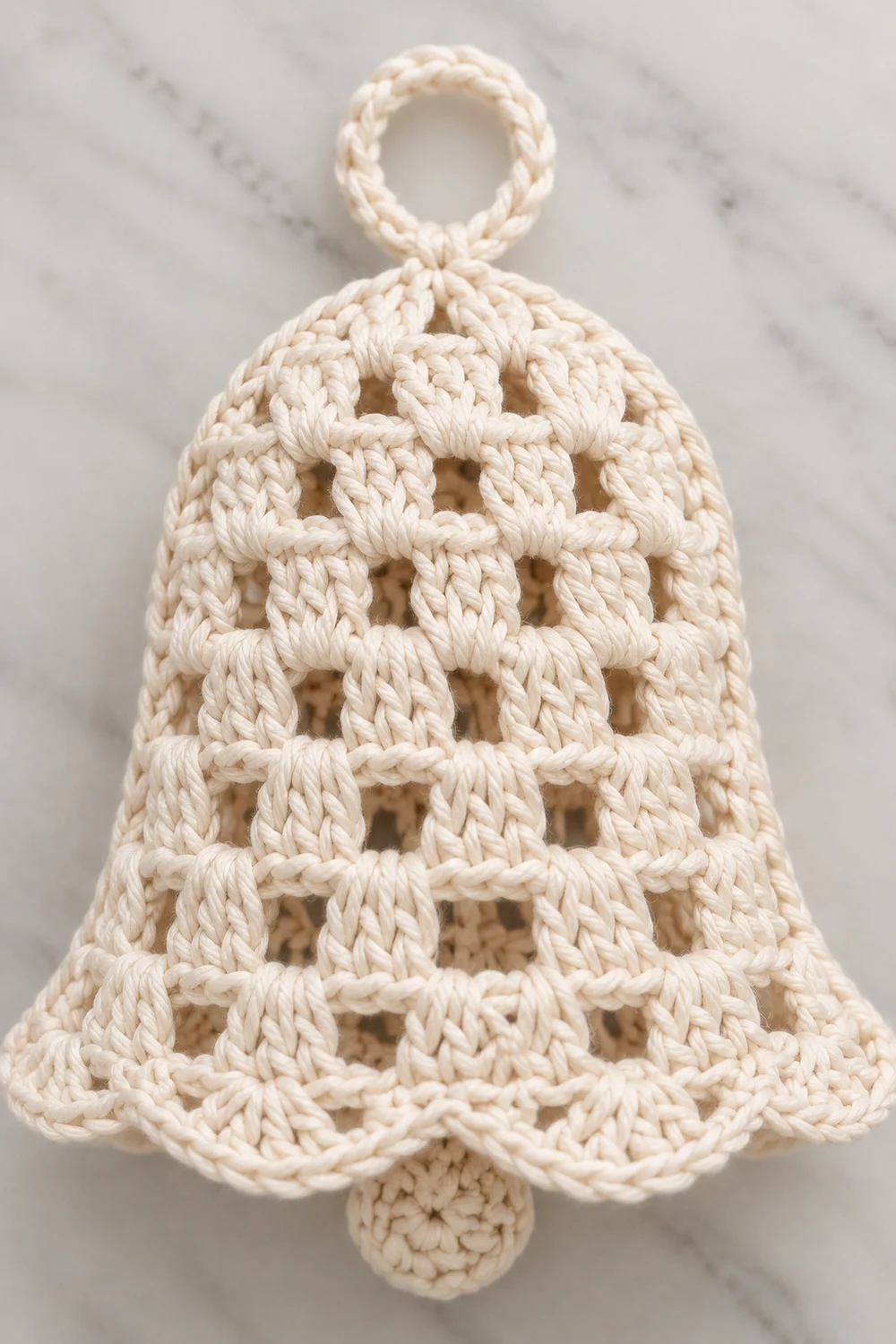 Cream-colored crocheted bell ornament with open lattice pattern and hanging loop on marble surface.