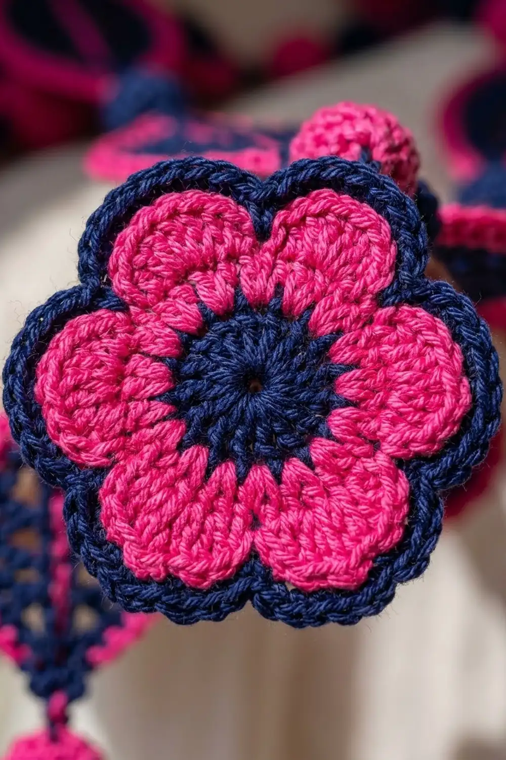 Handmade crocheted flower featuring bright pink textured petals surrounding a dark navy center, with more flowers softly blurred behind.