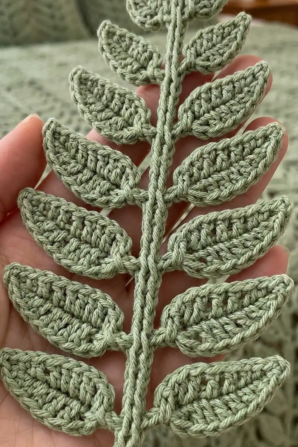 Crocheted pale green fern-like plant with textured leaf stitching held in someone's palm against soft background.