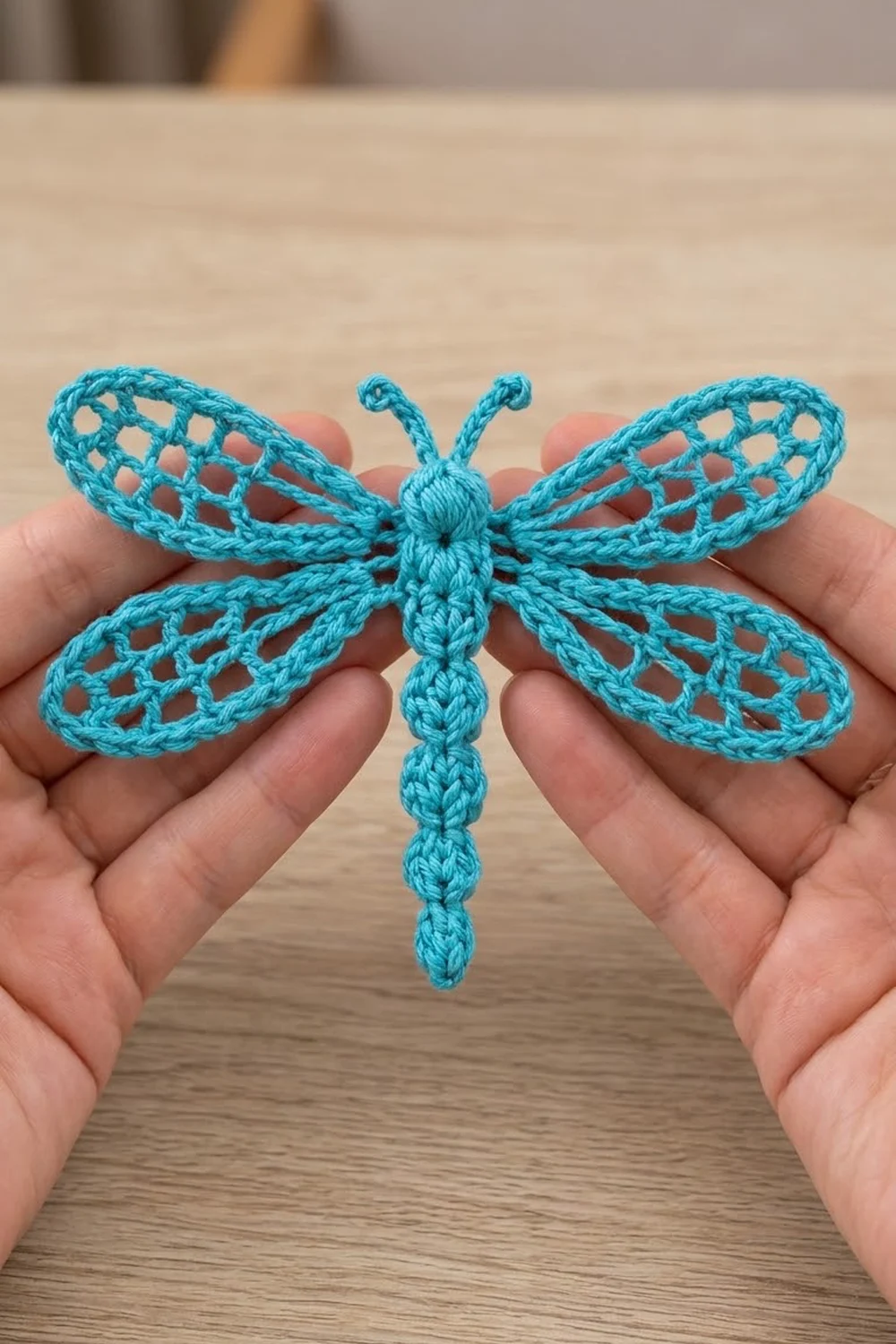 A bright turquoise crocheted dragonfly with intricate lace wings is carefully held in someone's hands.