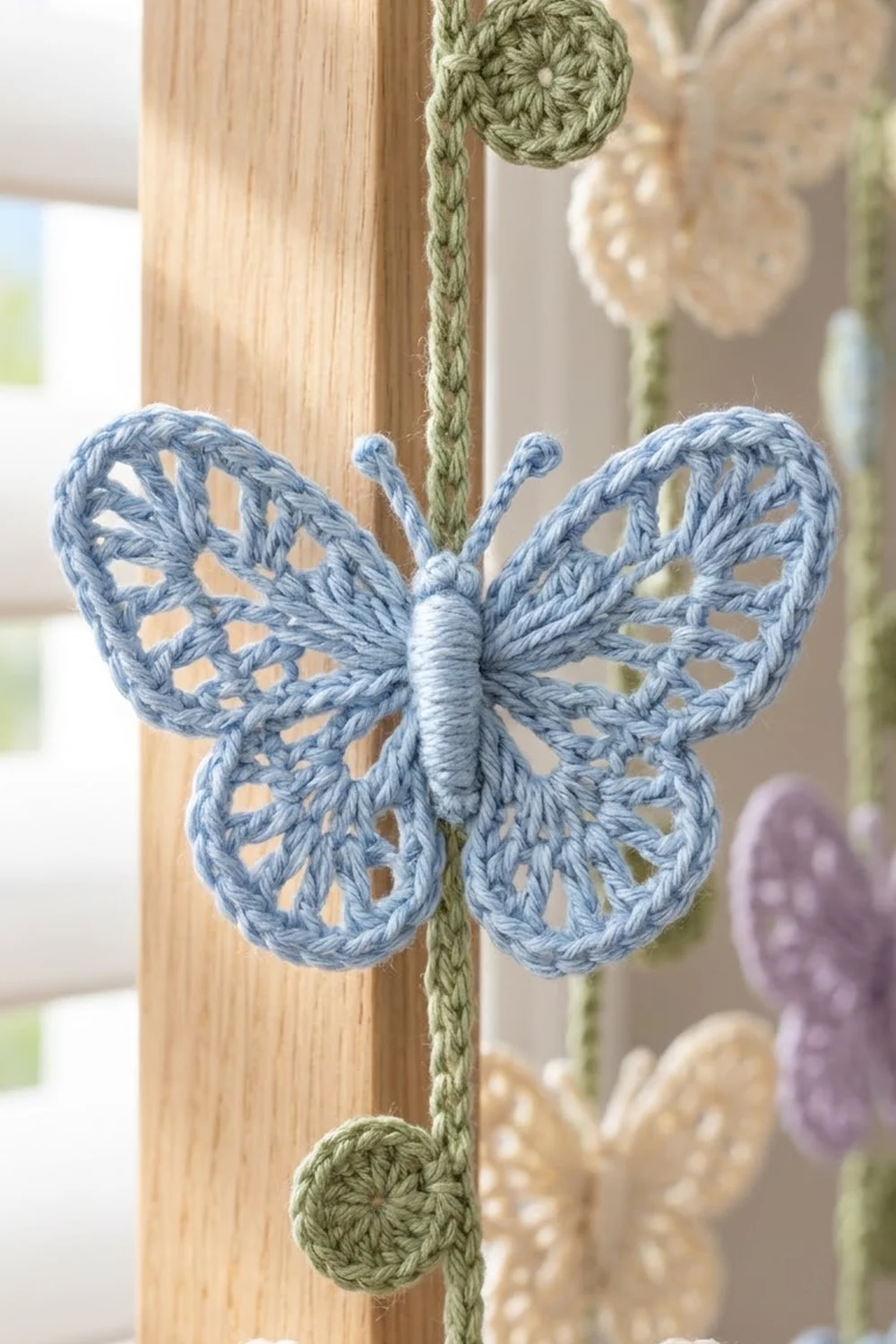 Blue crocheted butterfly with delicate openwork wings hangs from a green cord on natural wood, with colorful ornaments softly blurred behind.