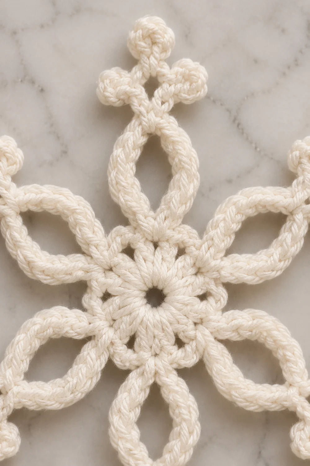 Close-up of a cream-colored crocheted snowflake ornament with intricate knotted details on a marble surface.