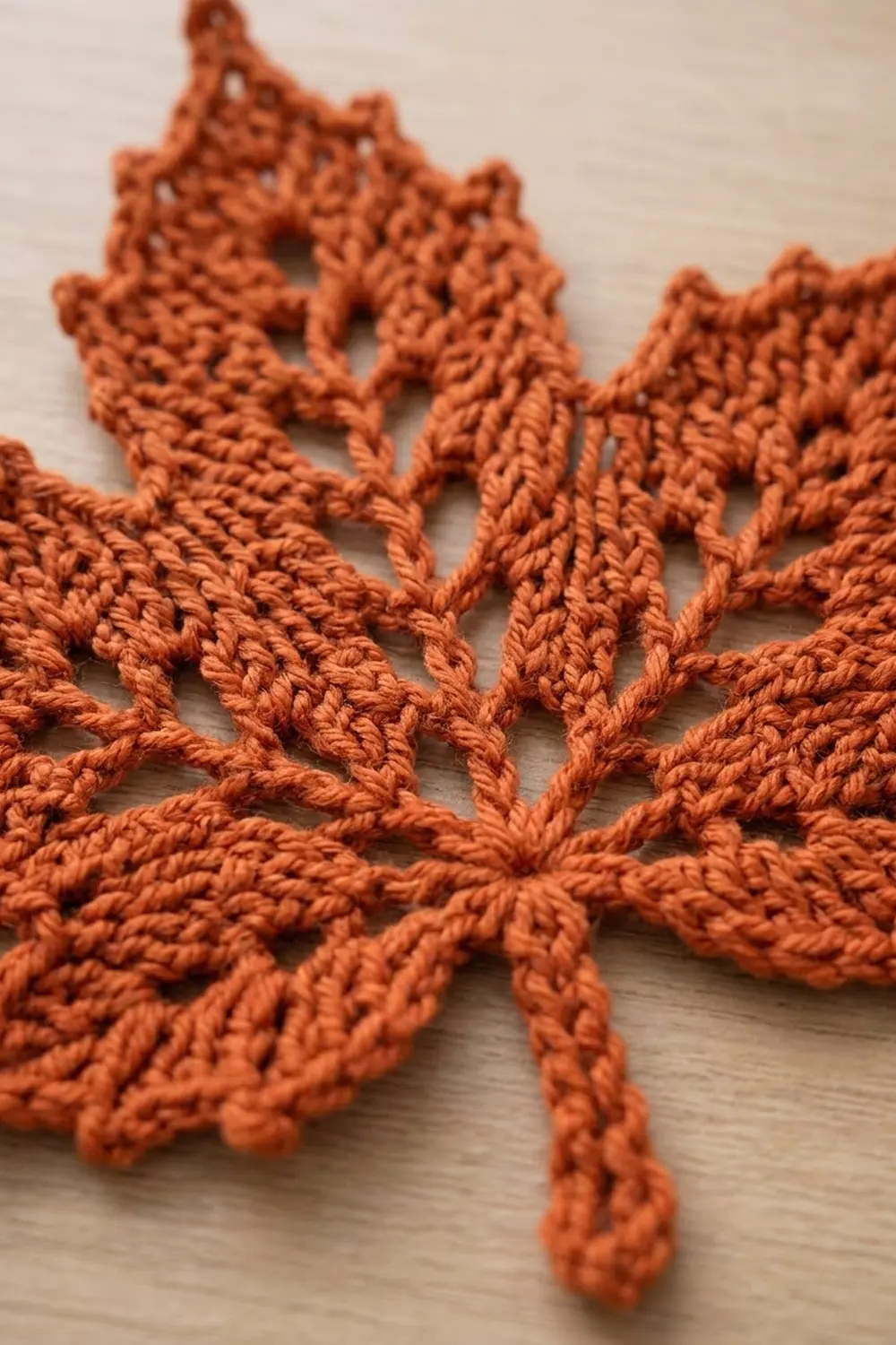 Close-up of intricate rust-colored crocheted snowflake with open lacework pattern on neutral wooden surface.