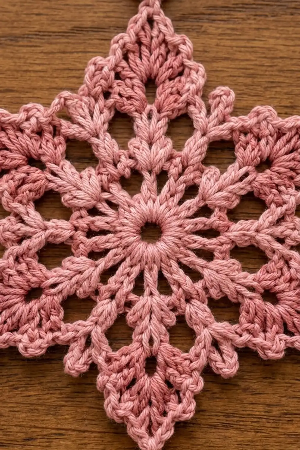 Delicate dusty rose crochet snowflake ornament with intricate lace pattern displayed on warm wood surface.