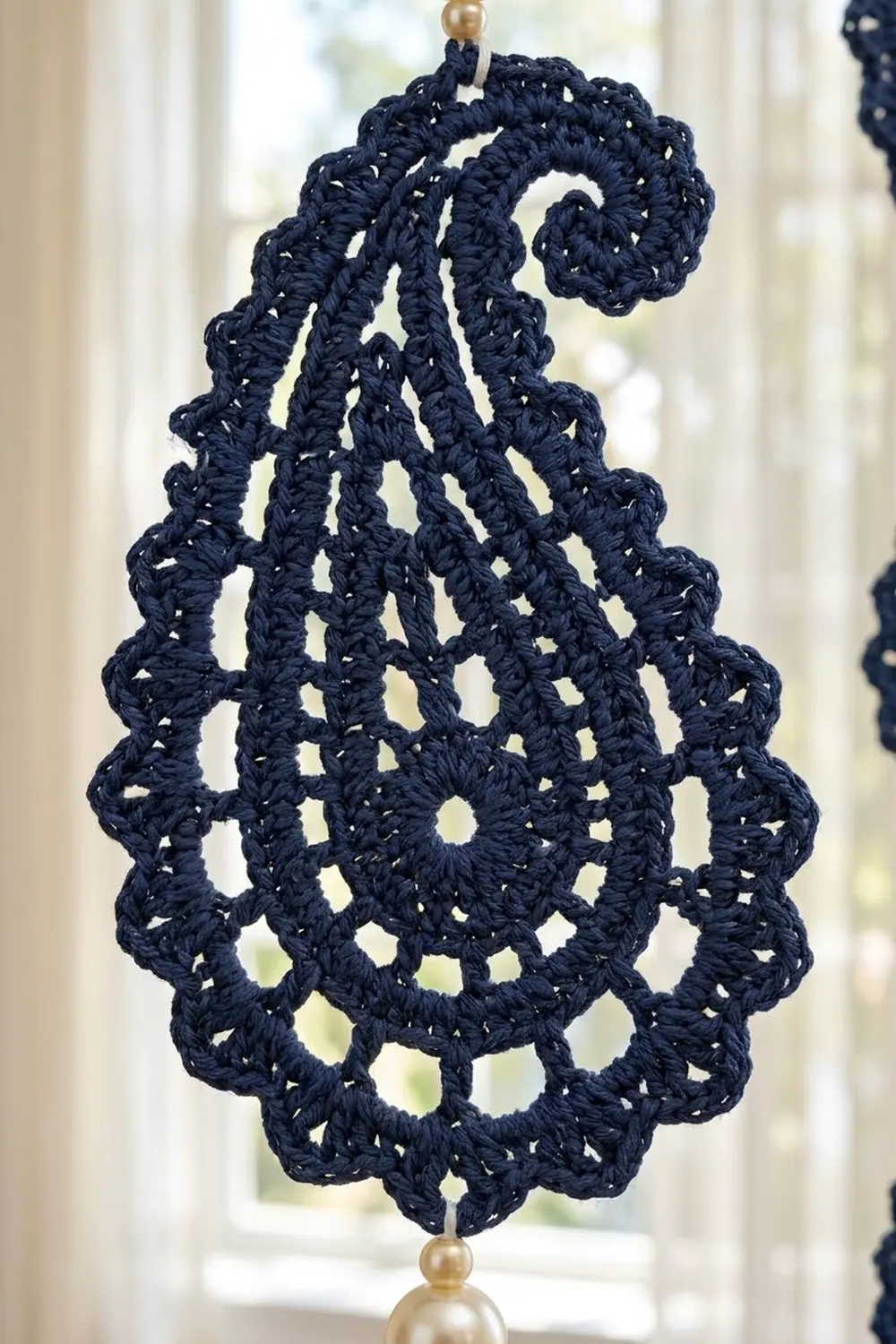 Dark navy crocheted teardrop ornament with openwork pattern and brass bead details, hanging in a window.