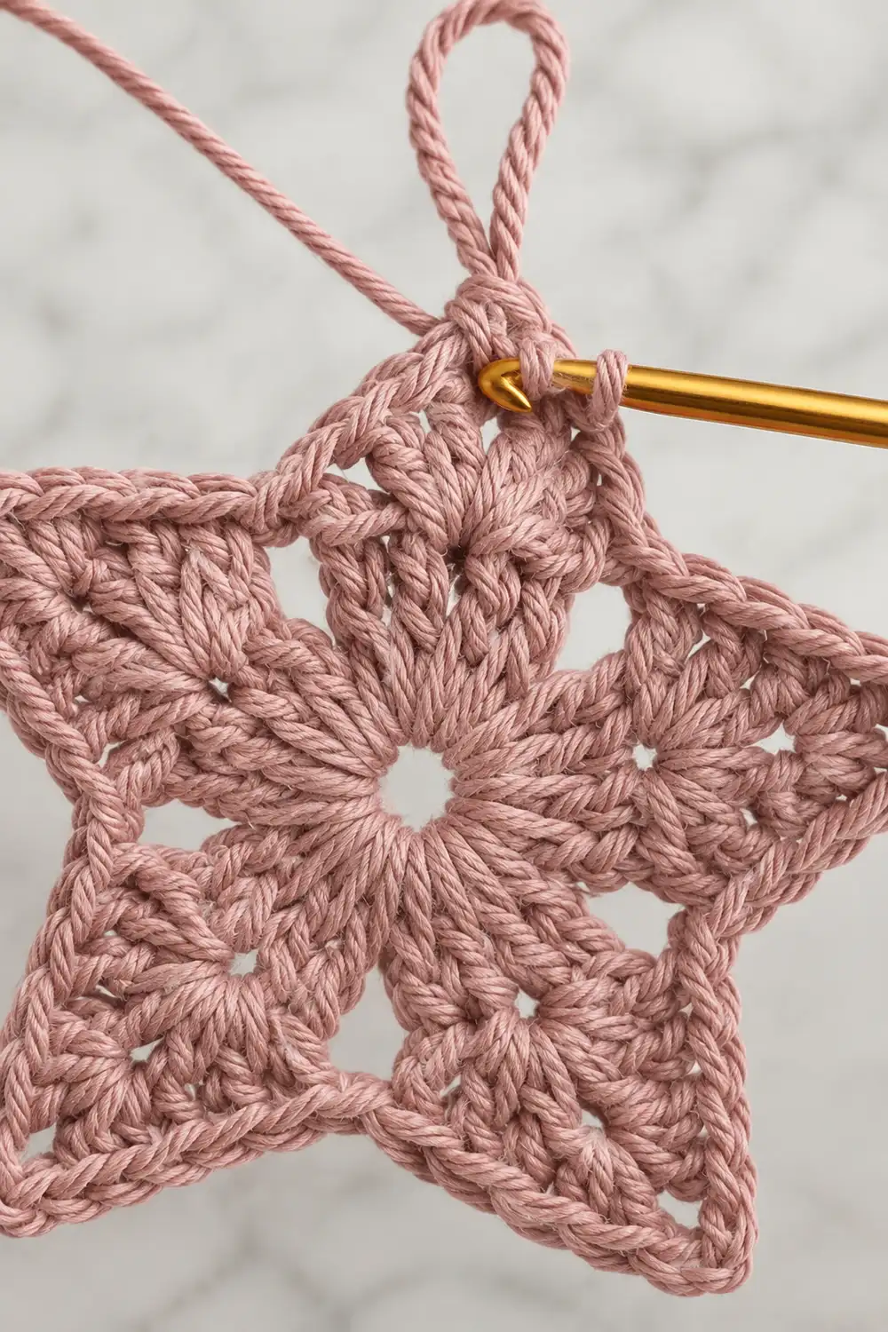 A pink crocheted star ornament with a loop and gold crochet hook on a white marble background.