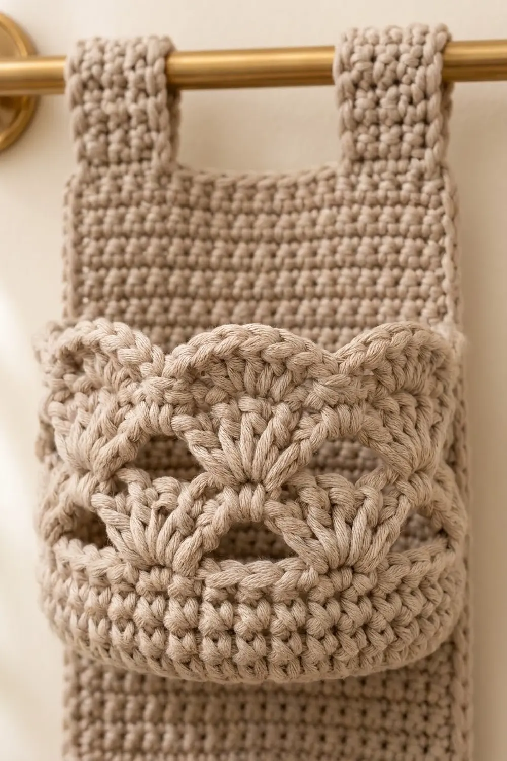 Closeup of cream-colored crocheted bag with braided bow detail hanging from a gold curtain rod against a soft beige background.
