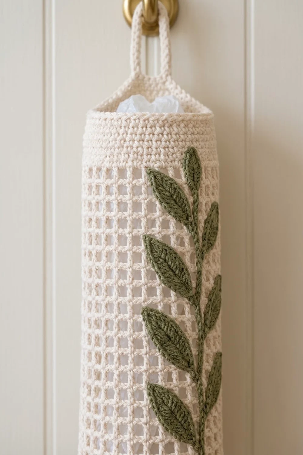 White crocheted hanging basket with green leaf appliqués on a brass hook against cream wall.