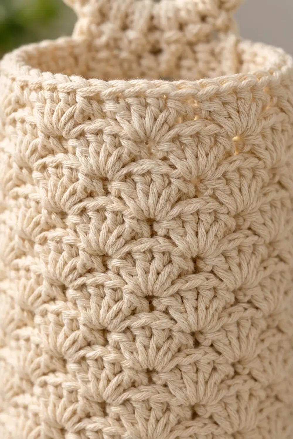 Close-up of cream-colored crocheted fabric showing detailed stitch texture with blurred green and beige background.