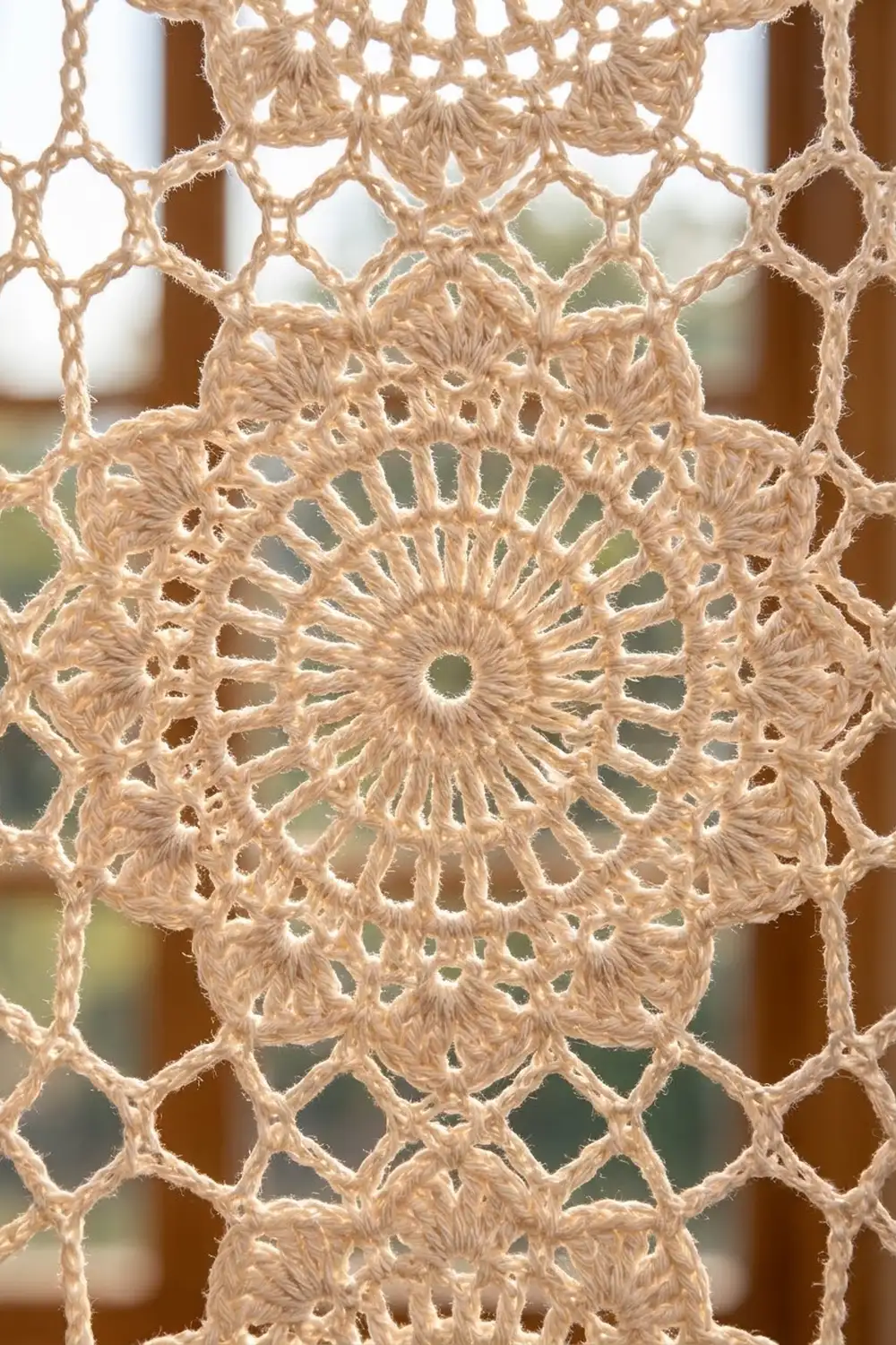 Tan crocheted doily with radiating geometric patterns, backlit by a window revealing soft blurred background through the lacework.
