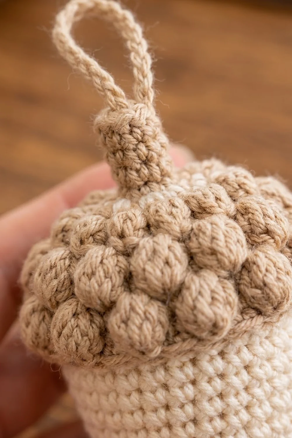 Beige crocheted pear with textured stitching and twisted stem held in a hand against a warm brown background.