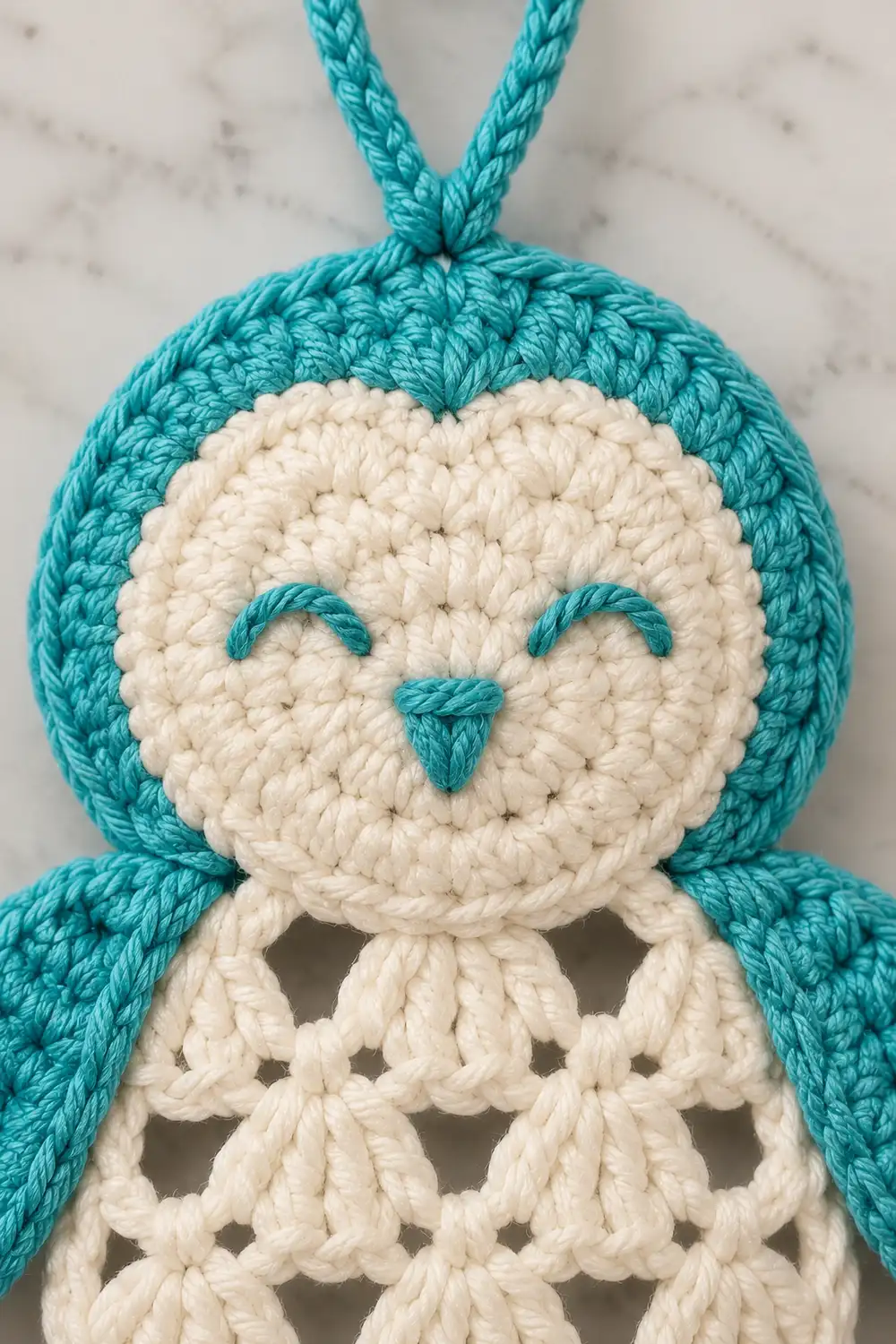 Crocheted owl ornament with turquoise body, cream face, and embroidered features hanging against marble background.