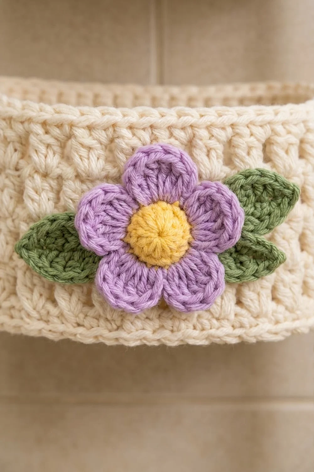 A purple crocheted flower with yellow center and green leaves appliquéd on cream-colored knitted fabric with textured ribbing.